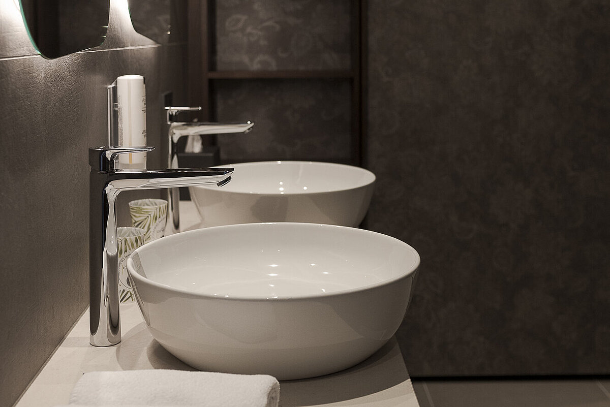 Modernes Badezimmer mit zwei weißen Waschbecken auf einer hellen Arbeitsplatte. Daneben sind zwei Handtücher ordentlich gefaltet. Ein großer Spiegel mit Beleuchtung hängt an der Wand, die aus einem dunklen Holzmaterial besteht.