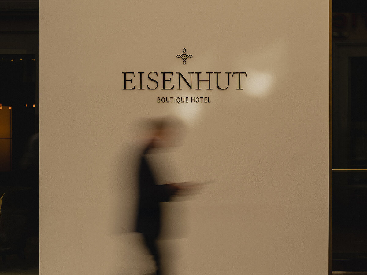 Eine verschwommene Silhouette einer Person läuft vor einer cremefarbenen Wand mit dem Schriftzug "Eisenhut Boutique Hotel".