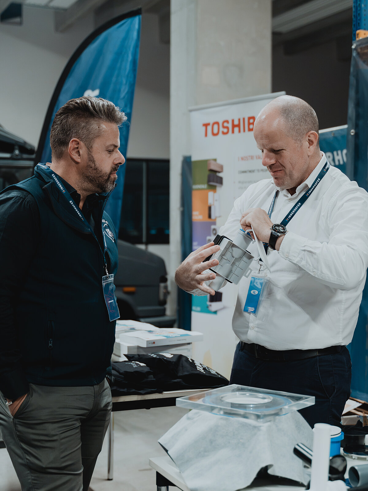 Zwei Männer stehen in einem Geschäft oder einer Ausstellung, einer hält ein technisches Gerät und zeigt es dem anderen. Im Hintergrund ist ein Toshiba-Banner zu sehen. Beide tragen Ausweise um den Hals.