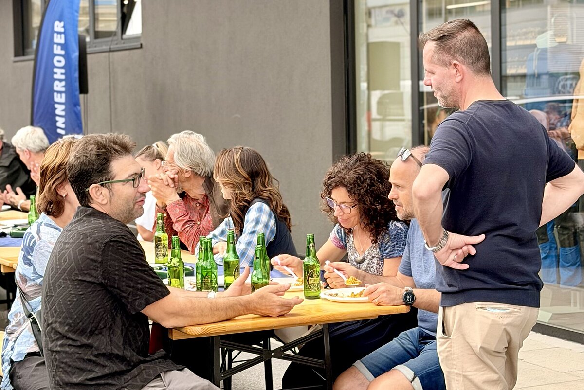 Gruppe von Menschen sitzt an einem Holztisch im Freien und unterhält sich, während sie Getränke genießen. Ein Mann steht daneben und unterhält sich mit der Gruppe. Die Szene spielt vor einem modernen Gebäude.