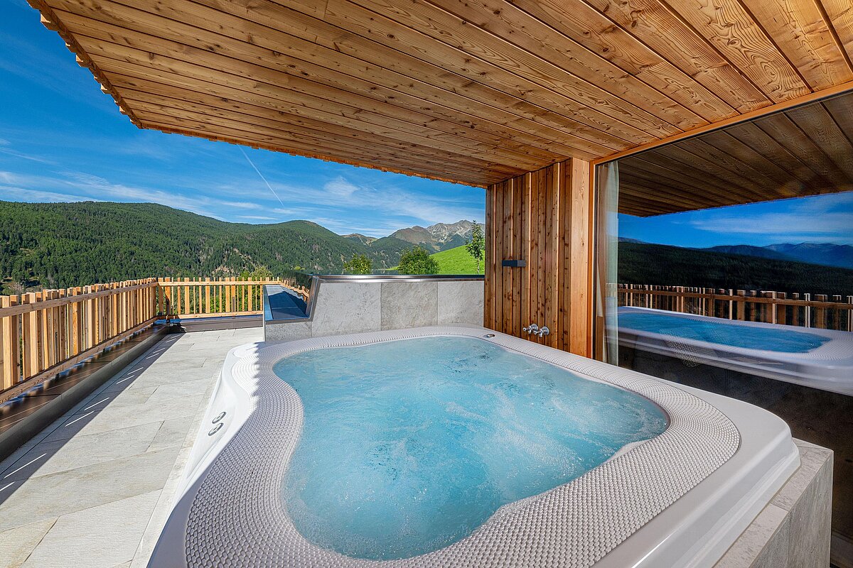 Whirlpool auf einer Terrasse mit Holzüberdachung, umgeben von Bergen und Wäldern. Die Umgebung ist sonnig mit klarem Himmel, und das Wasser im Whirlpool ist sprudelnd. Eine Glaswand reflektiert die Umgebung.