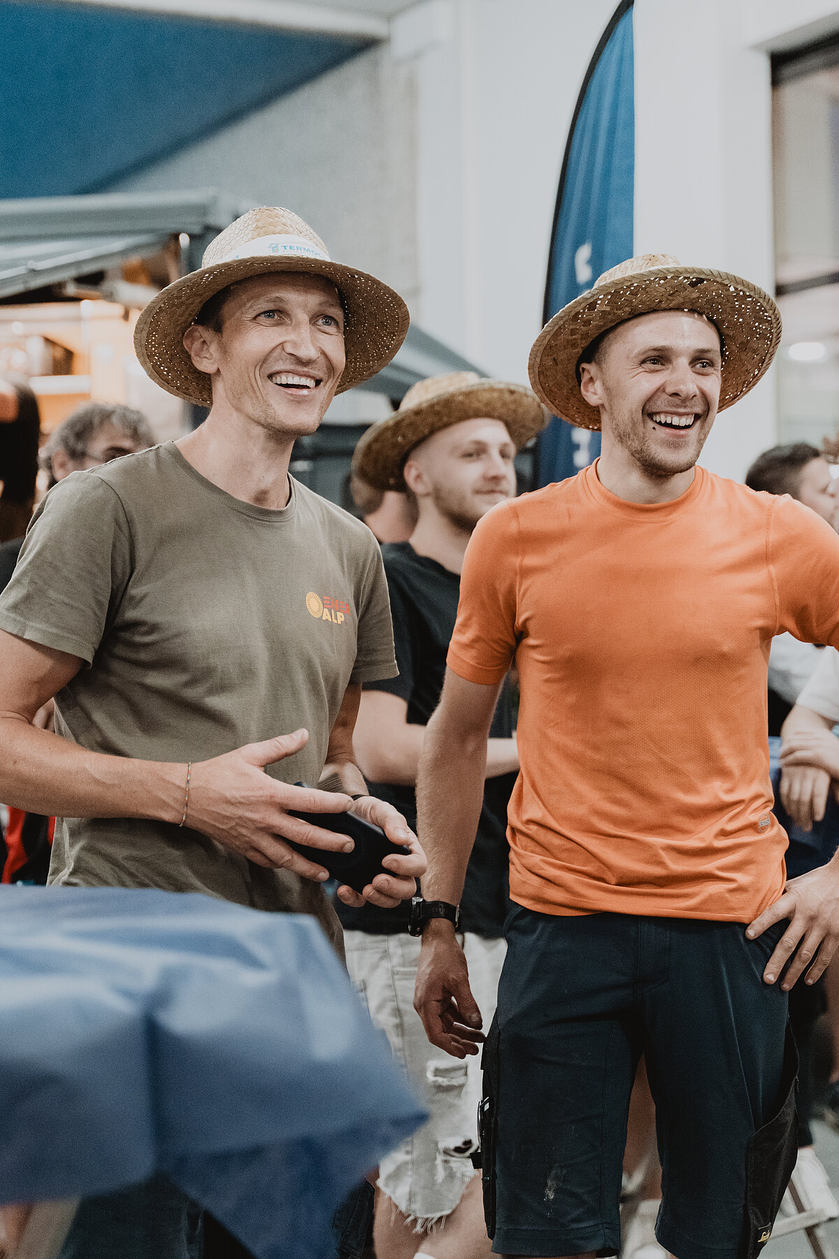 Zwei lachende Männer in Strohhüten stehen in einer Gruppe von Menschen. Der Mann links trägt ein olivgrünes T-Shirt, der rechts ein oranges. Im Hintergrund sind weitere Personen und ein blauer Banner zu sehen. Es scheint ein zwangloses, fröhliches Event zu sein.