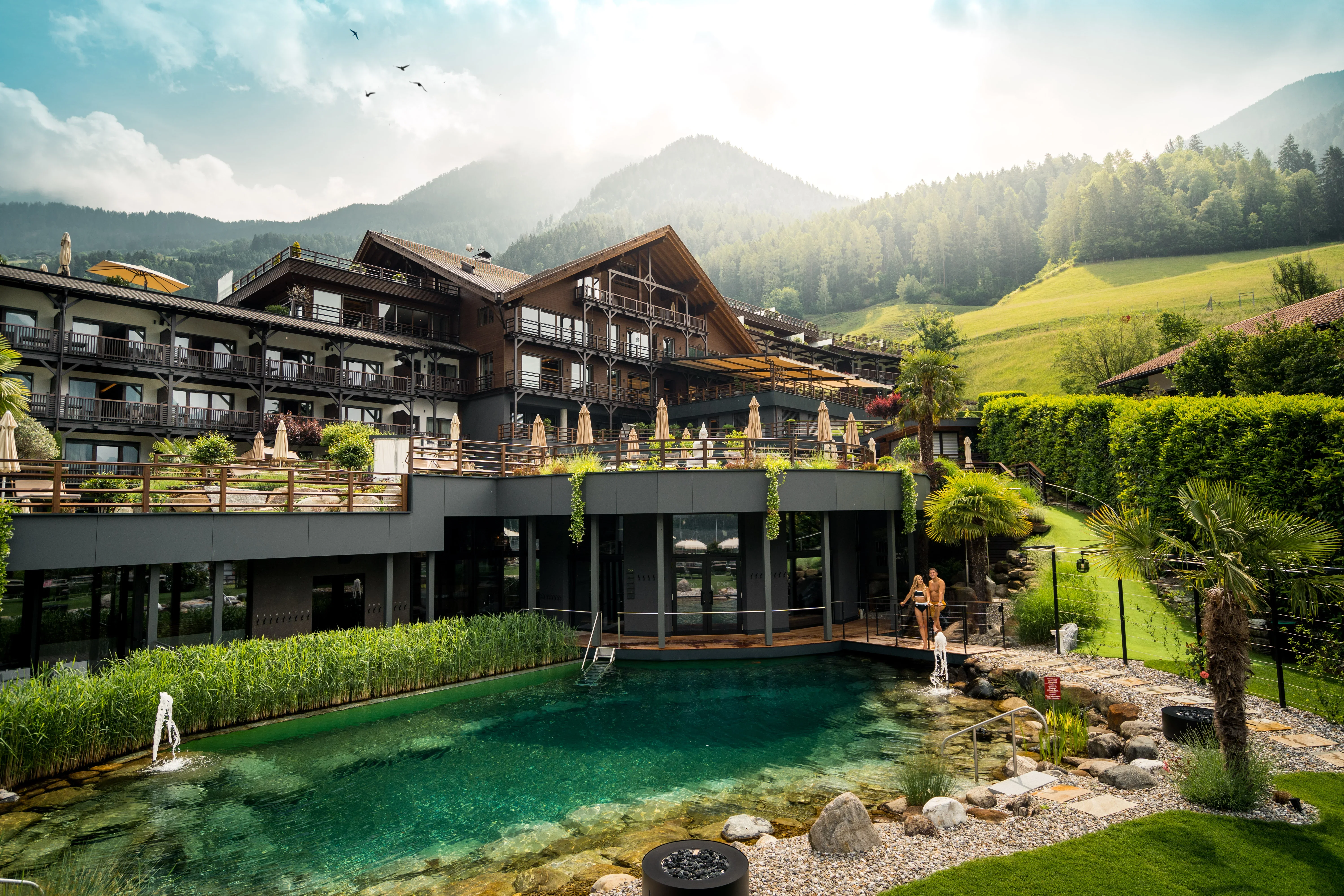 Luxuriöses Hotel mit mehreren Etagen und Balkonen, umgeben von grünen Hügeln und Bergen. Im Vordergrund ein großer, natürlicher Pool mit Pflanzen und Felsen. Der Himmel ist teilweise bewölkt, die Sonne scheint durch. Vögel sind am Himmel zu sehen.