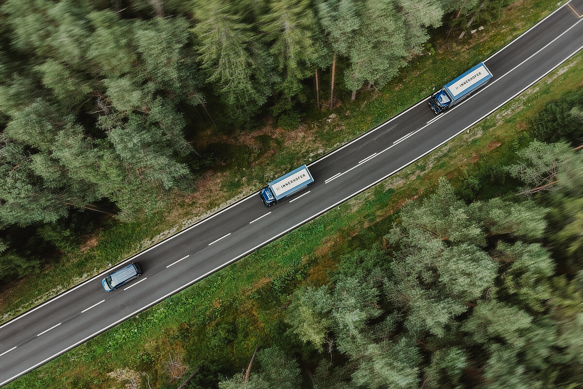 Drei blaue Lastwagen mit "IVECO" Aufschrift fahren hintereinander auf einer kurvenreichen Straße, die von dichtem Wald umgeben ist. Die Perspektive ist eine Luftaufnahme, die den grünen, bewaldeten Hintergrund betont.