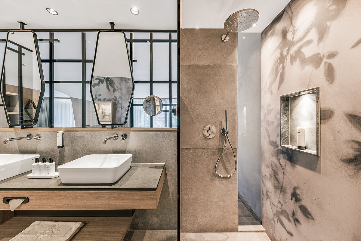 Modernes Badezimmer mit zwei rechteckigen Waschbecken auf einer Holzkonsole, dualen Wandspiegeln und eingebauter Regendusche. Die Wände sind in Erdtönen gehalten, die Beleuchtung ist dezent. Dekorative, schwarze Metallgitter an der Spiegelwand verleihen dem Raum einen eleganten Touch.