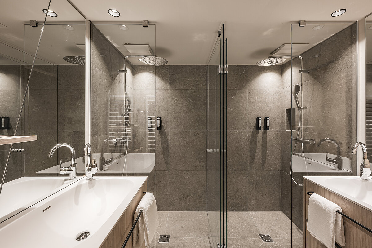 Modernes Badezimmer mit zwei Waschbecken, einem Glas-Duschbereich und großer Spiegelwand. Graue Fliesen dominieren das Design. Handtücher hängen an den Waschbecken. Dezente Deckenbeleuchtung sorgt für eine helle und elegante Atmosphäre. Zwei gleichmäßige Duschköpfe sind an der Decke montiert.