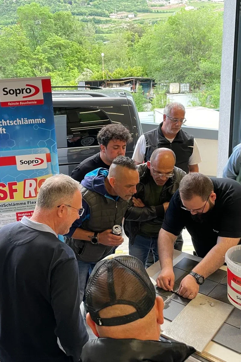 Eine Gruppe von Männern steht neugierig um einen Tisch versammelt, während einer von ihnen eine Kachel demonstriert oder repariert. Im Hintergrund ist ein Werbeplakat für "Sopro DSF" zu sehen. Die Szene findet anscheinend in einer Werkstatt oder einem Arbeitsraum statt.