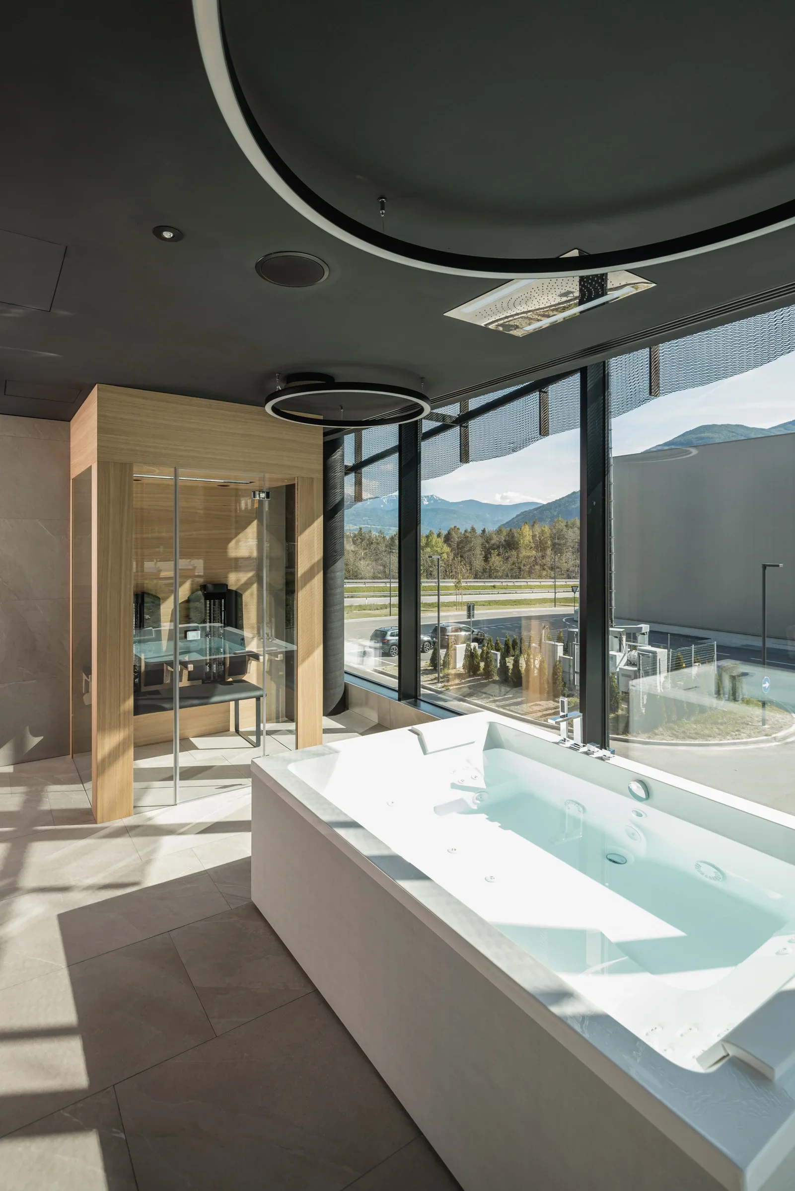 Modernes Badezimmer mit großer Badewanne und Sauna, großer Fensterfront mit Blick auf die Landschaft und Berge im Hintergrund. Elegantes, minimalistisches Design mit dunkler Decke und hellen Fliesen.
