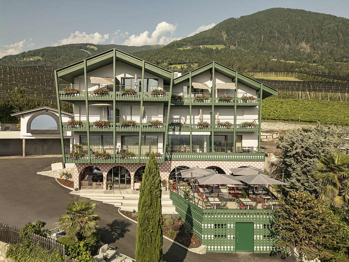 Ein mehrstöckiges Hotel im traditionellen alpinen Stil, mit Balkonen voller Blumen. Es steht vor einer hügeligen Landschaft mit Weinfeldern und Wäldern im Hintergrund. Eine Terrasse mit Sonnenschirmen ist auf der rechten Seite des Gebäudes sichtbar.