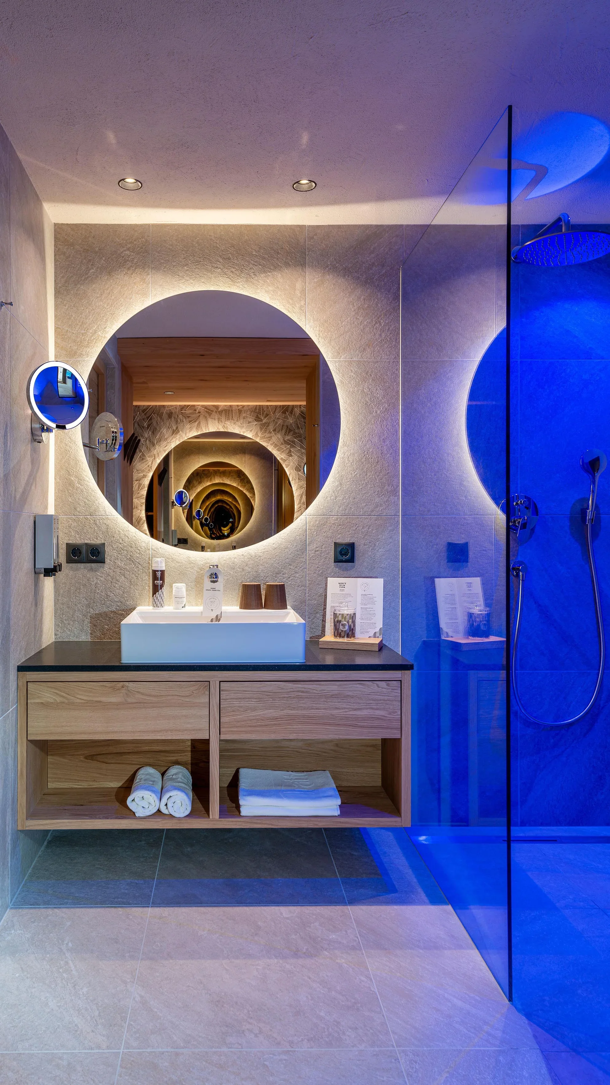 Modernes Badezimmer mit rundem Spiegel, hinterleuchtet und einem Waschbecken auf einem Holzunterschrank. Die Duschkabine rechts hat blaues Licht. Handtücher liegen ordentlich gefaltet im Regal unter dem Waschbecken. Elegantes Design mit Fokus auf Beleuchtung und Stil.