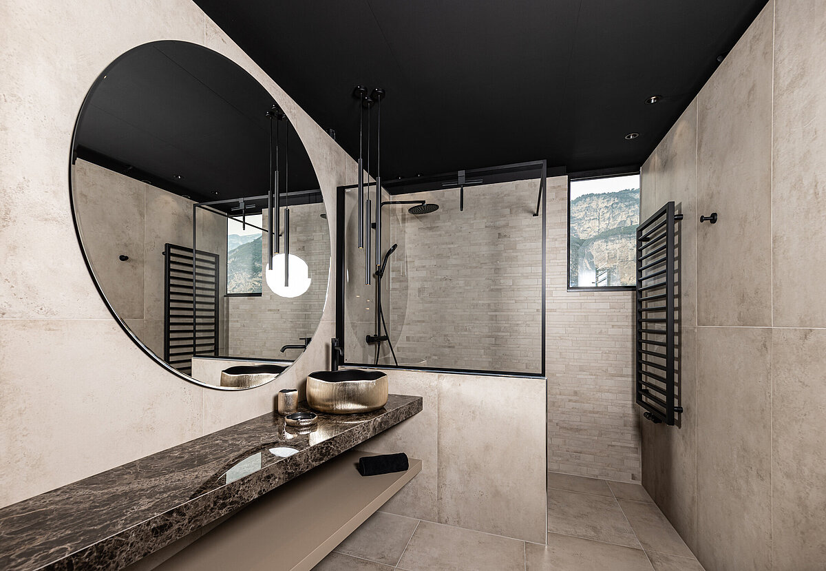 Modernes Badezimmer mit großen beige Fliesen, einem großen runden Spiegel, einem Waschbecken auf einer Marmorplatte und einer offenen Dusche. Ein Fenster bietet Blick auf eine Berglandschaft. Schwarzer Handtuchhalter und Armaturen ergänzen das Design.