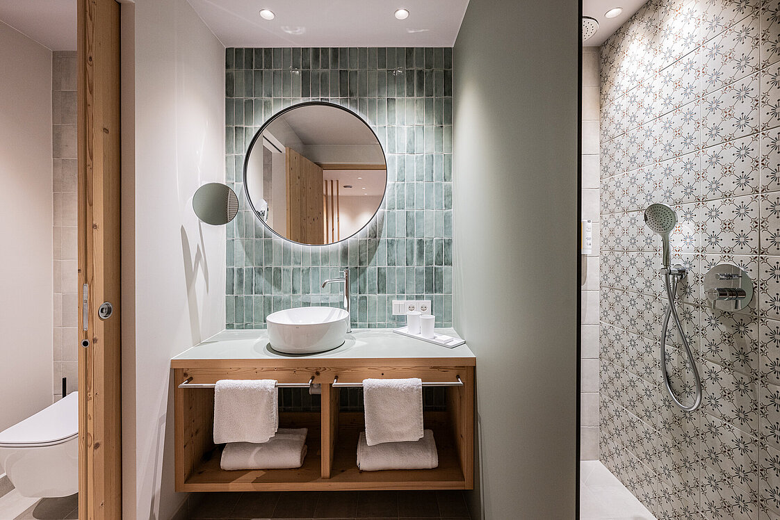Modernes Badezimmer mit großem runden Spiegel über einem Waschtisch mit einem Waschbecken. Unter dem Waschtisch sind weiße Handtücher gefaltet. Rechts daneben befindet sich eine Dusche mit gemusterten Fliesen. Auf der linken Seite führt eine Tür zu einer separaten Toilette.