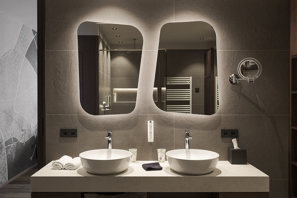 Modernes Badezimmer mit zwei runden Waschbecken auf einem weißen Doppelwaschtisch. Darüber hängen asymmetrische Spiegel, die weich beleuchtet sind. Daneben ein Vergrößerungsspiegel. Handtücher und Pflegeprodukte ergänzen die stilvolle Einrichtung.