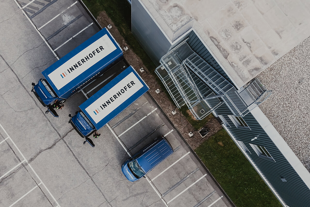 Zwei blaue LKW mit der Aufschrift "Innerhofer" parken parallel nebeneinander in einem leeren Parkplatz neben einem Gebäude. Ein blauer Lieferwagen steht daneben. Zwei Personen stehen bei den LKWs. Der Gebäudeeingang ist über eine Stahltreppe erreichbar. Luftaufnahme.