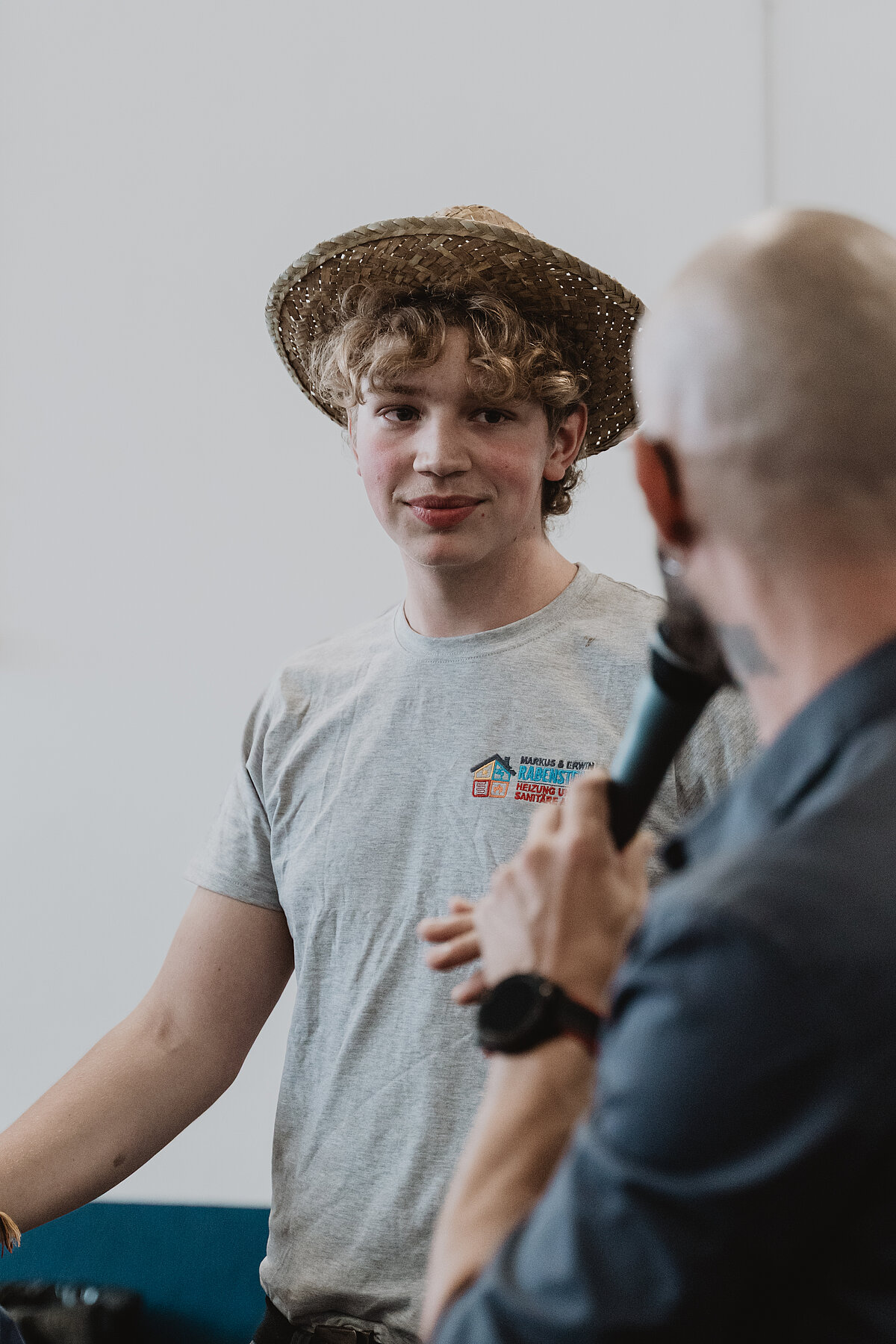 Ein junger Mann mit Locken trägt einen Strohhut und ein graues T-Shirt, während er einen älteren Mann anhört, der ein Mikrofon in der Hand hält. Sie befinden sich in einem Raum mit weißem Hintergrund.
