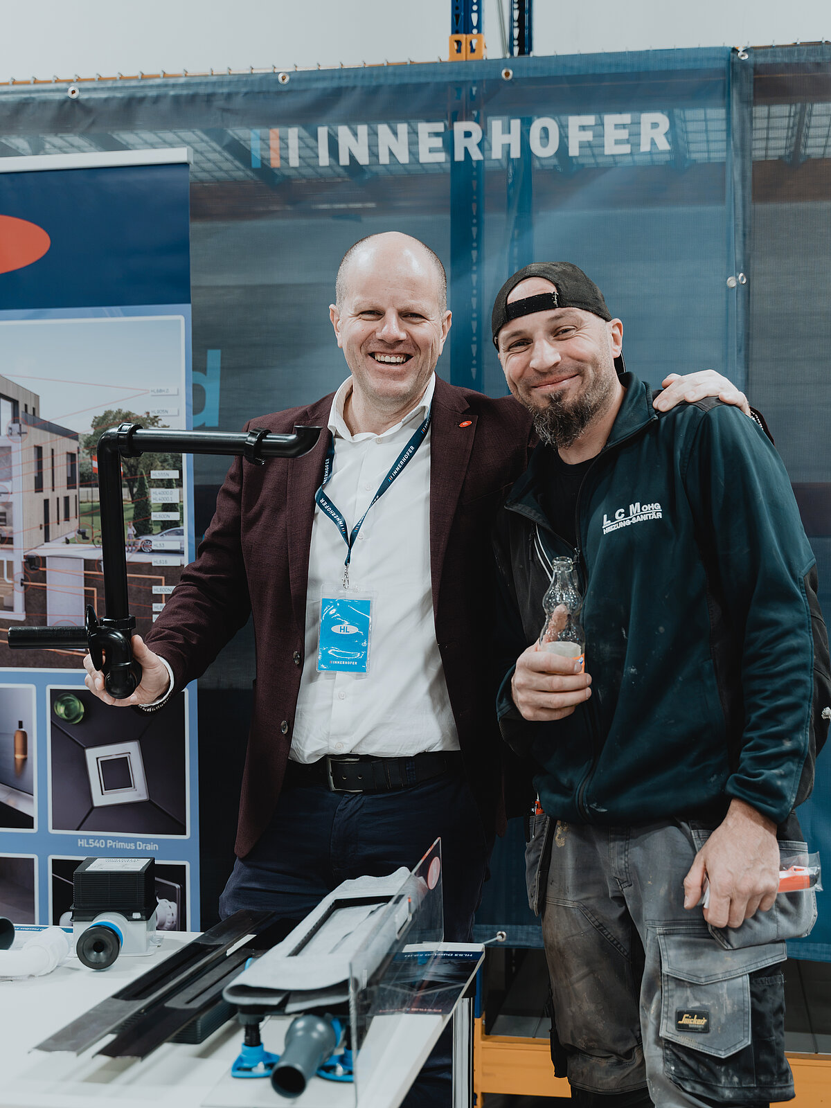 Zwei Männer stehen lächelnd nebeneinander auf einer Messe. Der eine trägt einen Anzug und hält ein Rohrstück, der andere ist leger gekleidet und hält eine Getränkeflasche. Im Hintergrund steht der Name "Innerhofer" auf einem Banner.