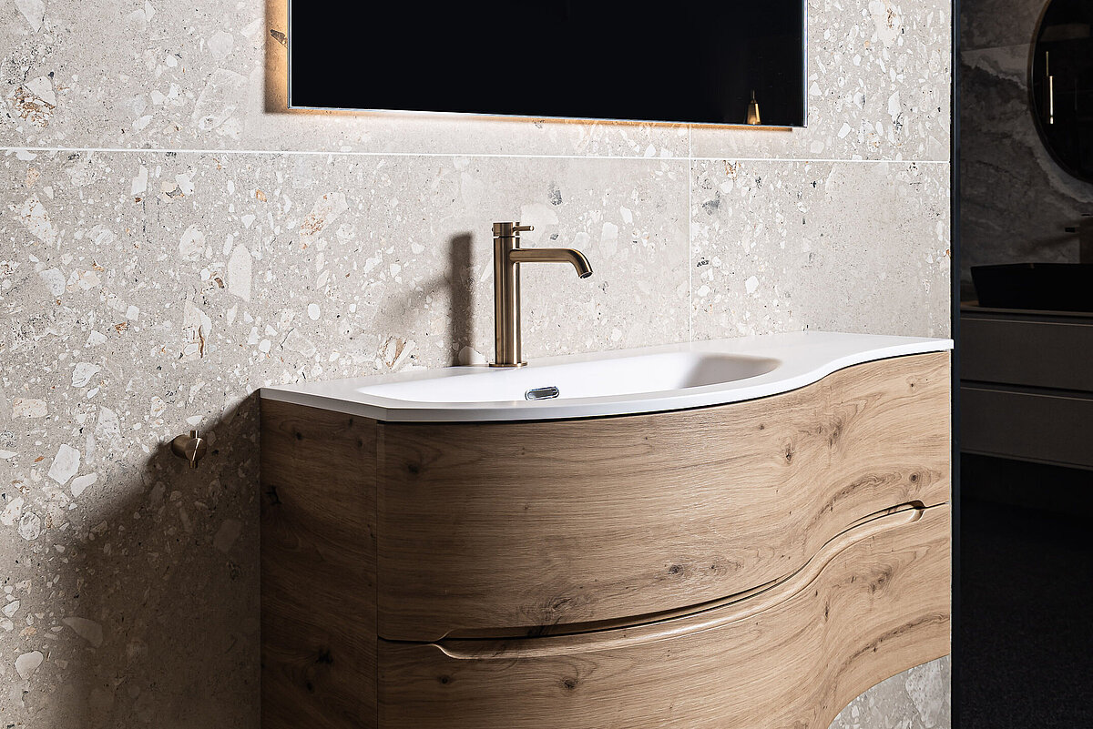 Ein modernes Badezimmer mit einem hängenden Waschbecken aus Holzoptik, eingebautem Wasserhahn und einem großen, rechteckigen Spiegel mit Beleuchtung darüber. Die Wand besteht aus hellgrauen Fliesen.