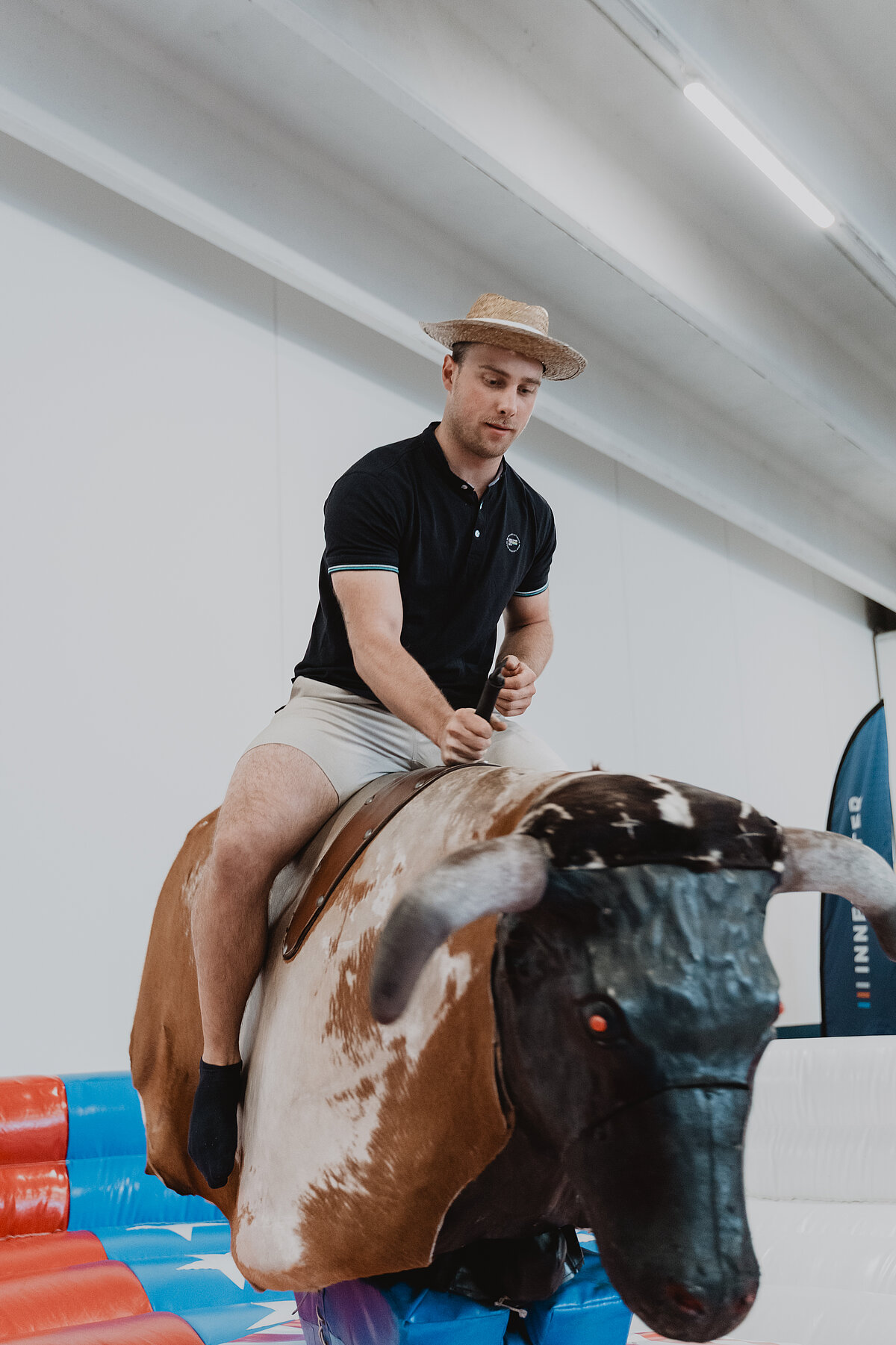 Ein Mann in kurzer Hose und Strohhut reitet auf einem mechanischen Bullen. Der Bulle steht auf einer bunten, aufblasbaren Unterlage in einem Innenraum. Der Mann hält sich am Griff des Bullen fest.