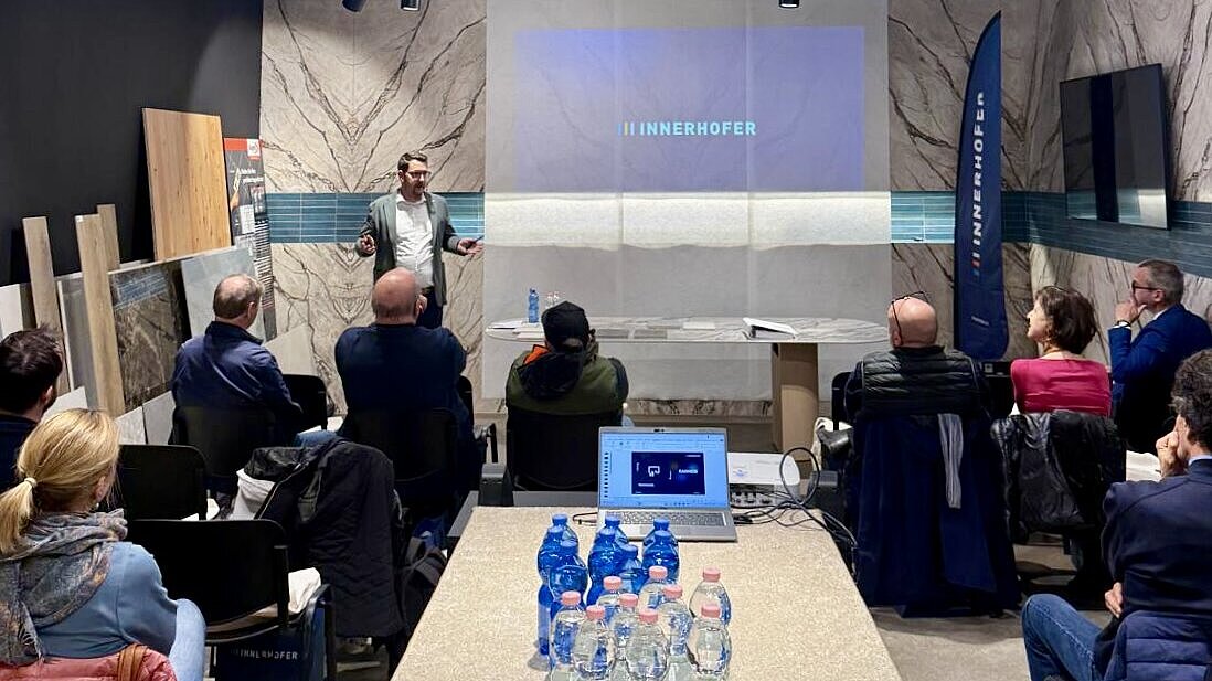 Eine Person hält eine Präsentation vor einer Gruppe von Zuhörern in einem modernen Konferenzraum. Auf dem Tisch im Vordergrund stehen Wasserflaschen. Die Wand im Hintergrund zeigt den Schriftzug "INNERHOFER".
