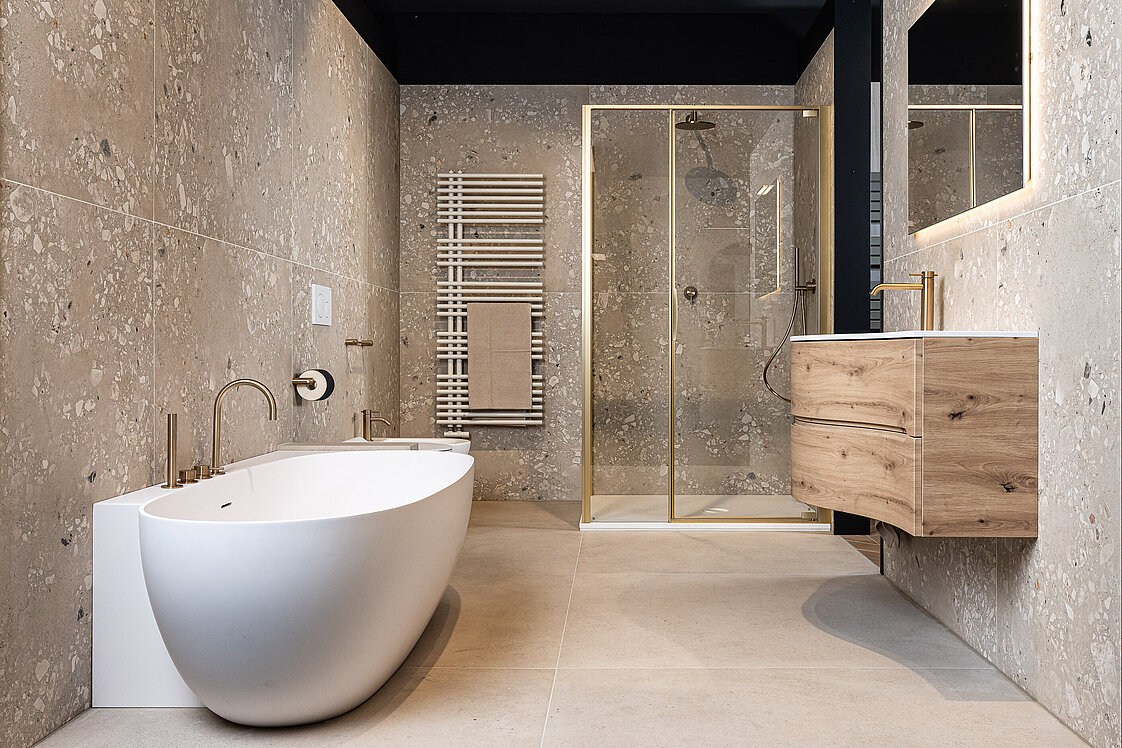 Modernes Badezimmer mit freistehender Badewanne, Regendusche mit Glastüren, Holzwaschtisch und rechteckigem Spiegel. Die Wände sind mit natursteinähnlichen Fliesen verkleidet und ein Handtuchheizkörper befindet sich neben der Dusche.