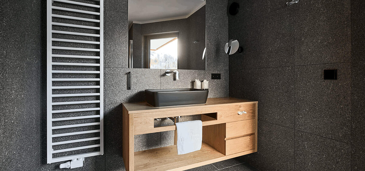 Modernes Badezimmer mit dunkler Steinwand, großem Spiegel, Holzschränken und Badewanne. Im Hintergrund ist ein Flur mit Tageslicht zu sehen.