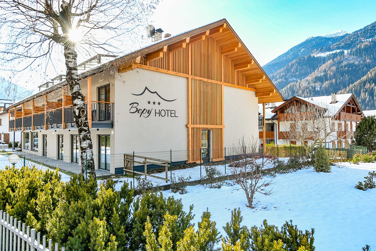 Ein modernes Hotelgebäude mit hölzernen Akzenten und einem Logo, umgeben von schneebedecktem Gelände und Bergen im Hintergrund. Ein Sonnenstrahl scheint durch die Bäume.