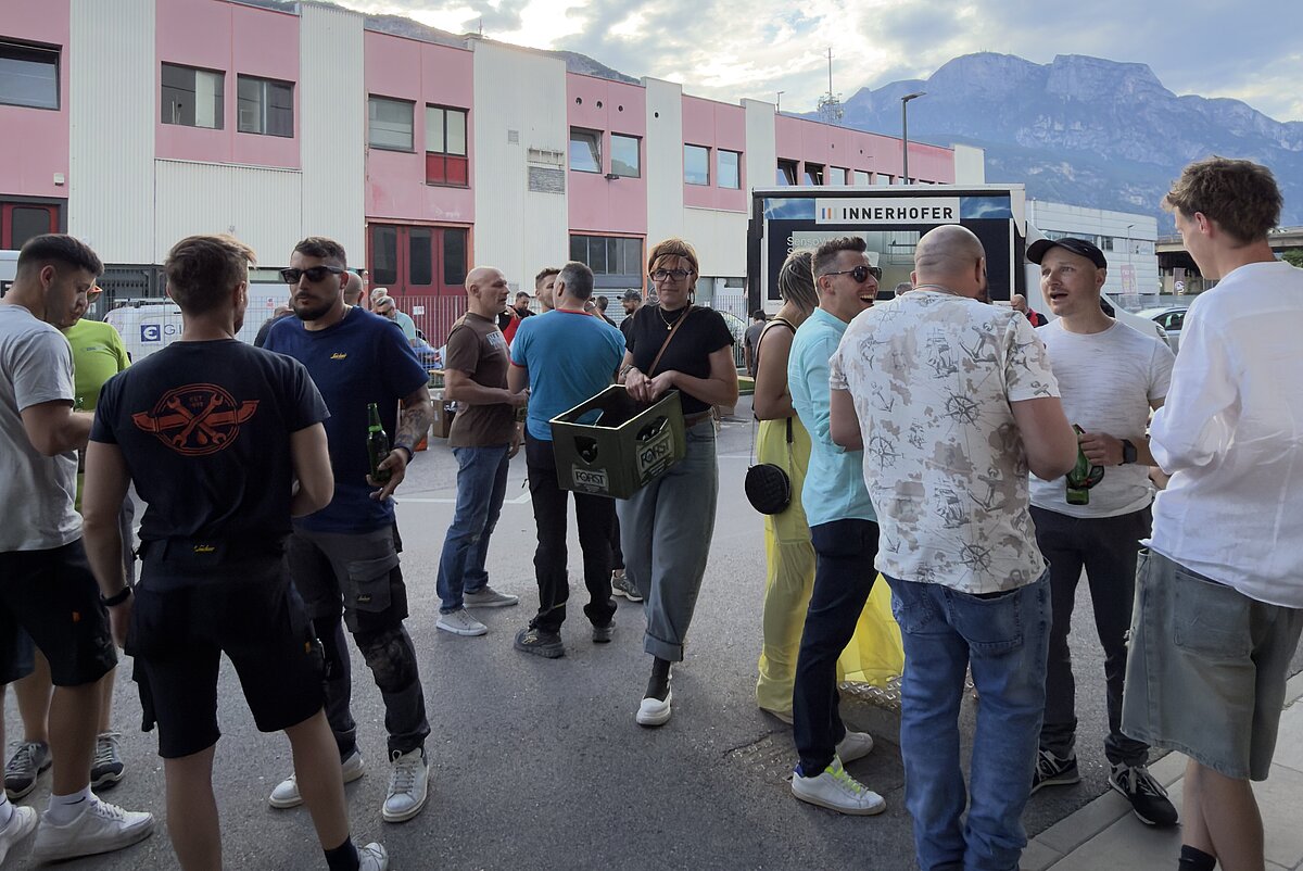 Eine Gruppe von Menschen steht im Freien auf einer Straße, offenbar bei einem sozialen Treffen. Im Hintergrund sind ein modernes Gebäude und eine hügelige Landschaft unter einem bewölkten Himmel zu sehen.
