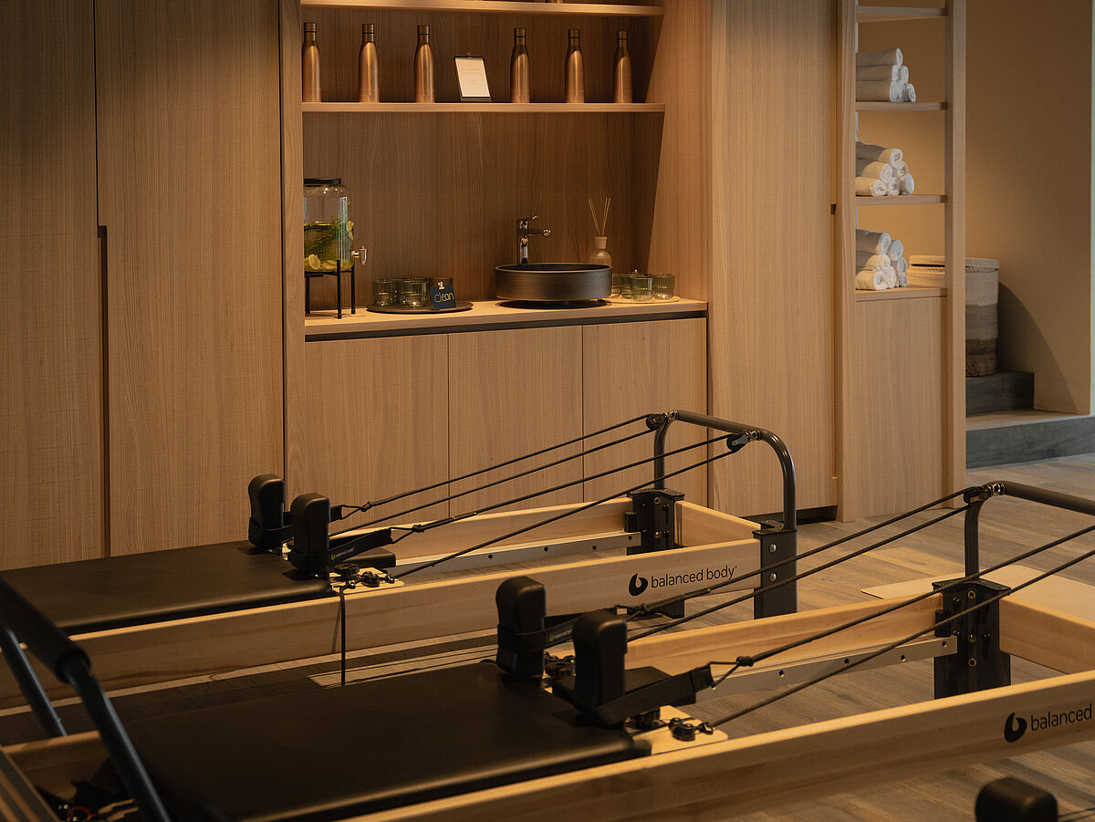 Ein moderner Pilatesraum mit Holzkonstruktion, offenen Regalen mit Dekoration und Flaschen, sowie mehreren Reformern im Vordergrund. Die Atmosphäre ist ruhig und minimalistisch.