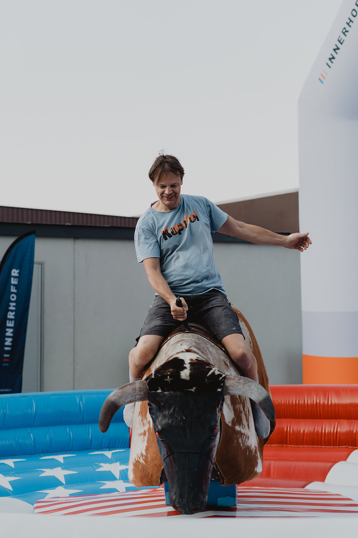 Ein Mann reitet auf einem mechanischen Bullen bei einer Outdoor-Veranstaltung. Er trägt ein hellblaues T-Shirt und sieht konzentriert aus. Im Hintergrund sind Werbebanner und eine aufblasbare Struktur erkennbar.