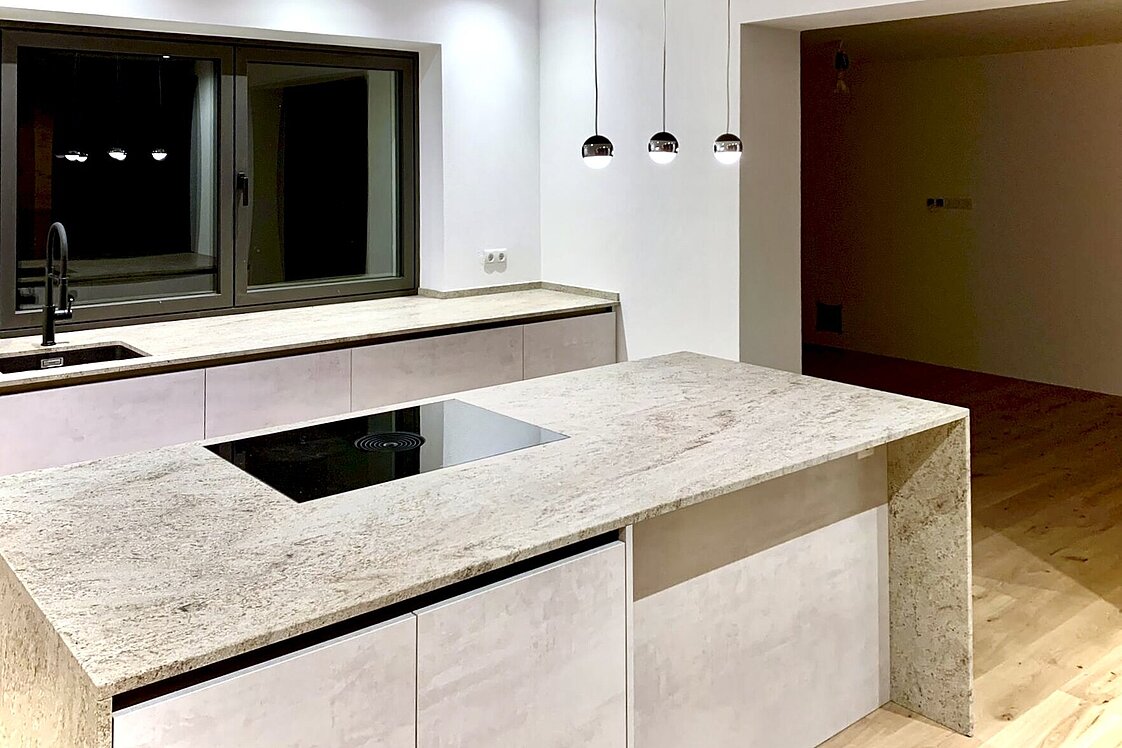 Moderne Küche mit heller Stein-Arbeitsplatte, Kochinsel und schwarzem Kochfeld. Hängende Pendelleuchten über der Insel. Große Fenster und minimalistisches Design. Holzfußboden und offene, helle Raumgestaltung.