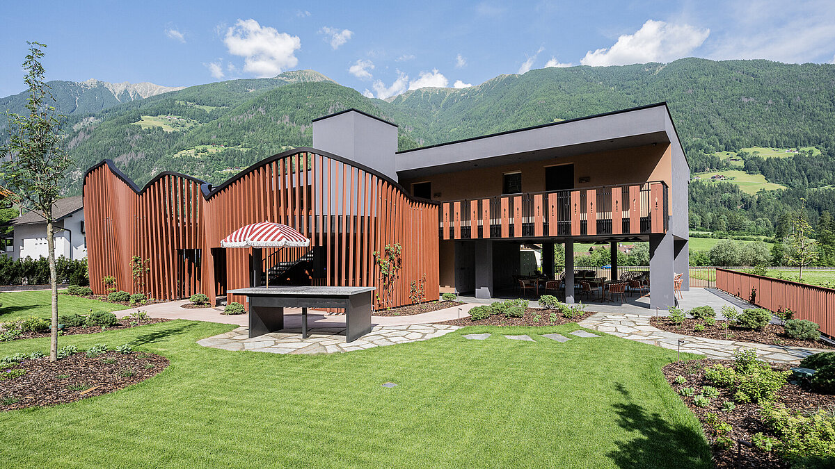 Modernes zweistöckiges Haus mit Holzverkleidung und Wellenarchitektur, umgeben von gepflegtem Garten. Vor dem Haus steht ein Tisch mit rotem Sonnenschirm. Im Hintergrund sind Berge und grünes Tal zu sehen, was eine idyllische Landschaft schafft.
