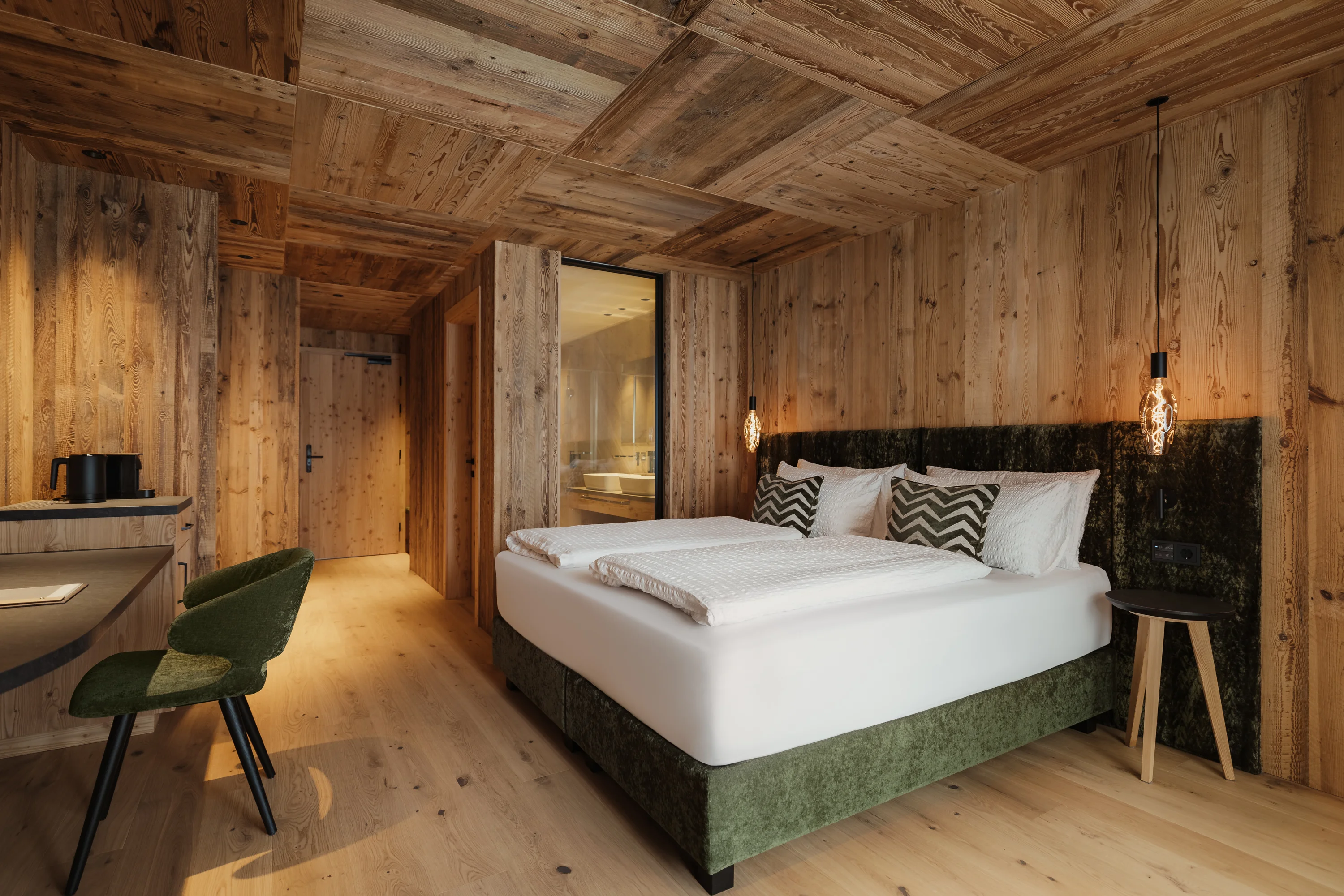 Ein stilvoll eingerichtetes Hotelzimmer mit Holzwänden und -decke. Es gibt ein großes Doppelbett mit Kissen, ein Schreibtisch mit grünem Stuhl, sowie dekorative Hängelampen. Der Raum wirkt warm und einladend.