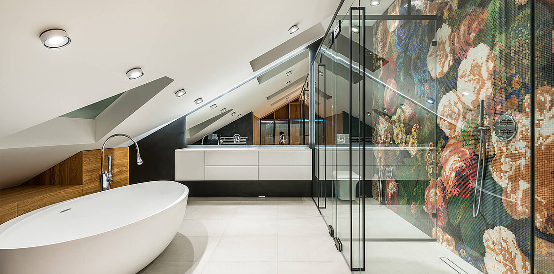 Ein modernes Badezimmer mit Dachschräge, freistehender Badewanne, großer Glasdusche und einem Blumenmuster an der Duschwand. Entlang der Rückwand befindet sich ein Doppelwaschtisch mit Spiegeln, alles in einem eleganten, minimalistischen Design.