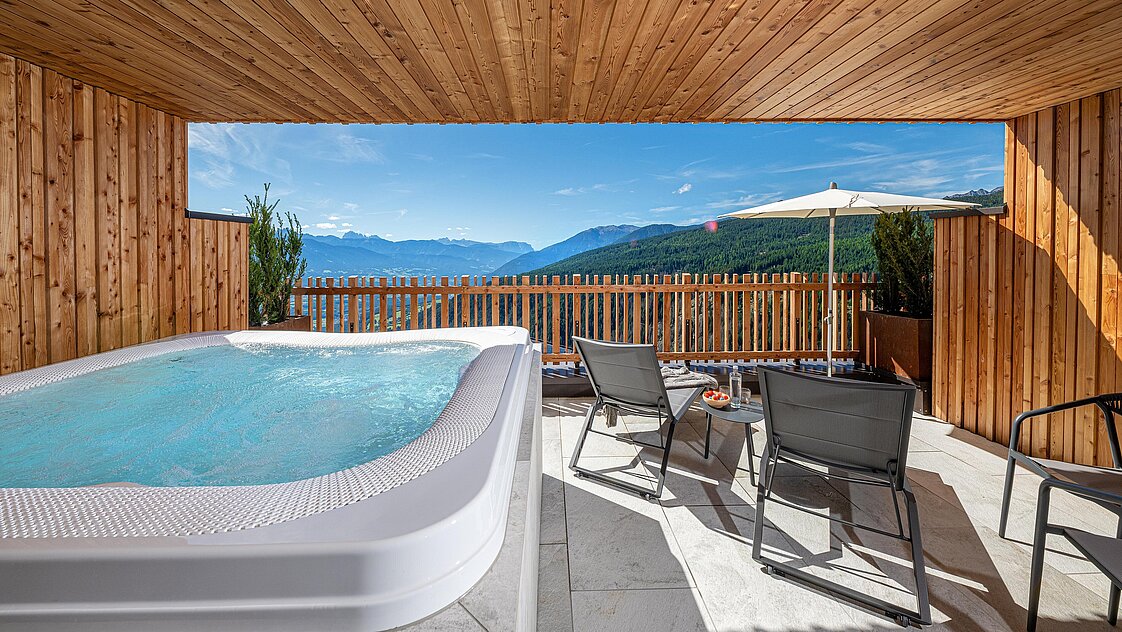 Holzverkleidete Terrasse mit einem Whirlpool, Blick auf die Berge und einen See. Sonnenschirme, Liegestühle und ein Tisch mit Getränken und Snacks komplettieren die entspannte Atmosphäre.