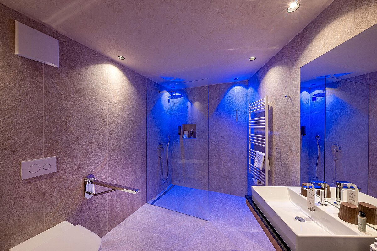 Modernes Badezimmer mit begehbarer Dusche, beleuchtet mit blauem Licht. An der Wand ein Handtuchhalter und ein Spiegel über einem Waschtisch mit Waschbecken. Dezente Spotbeleuchtung von der Decke.