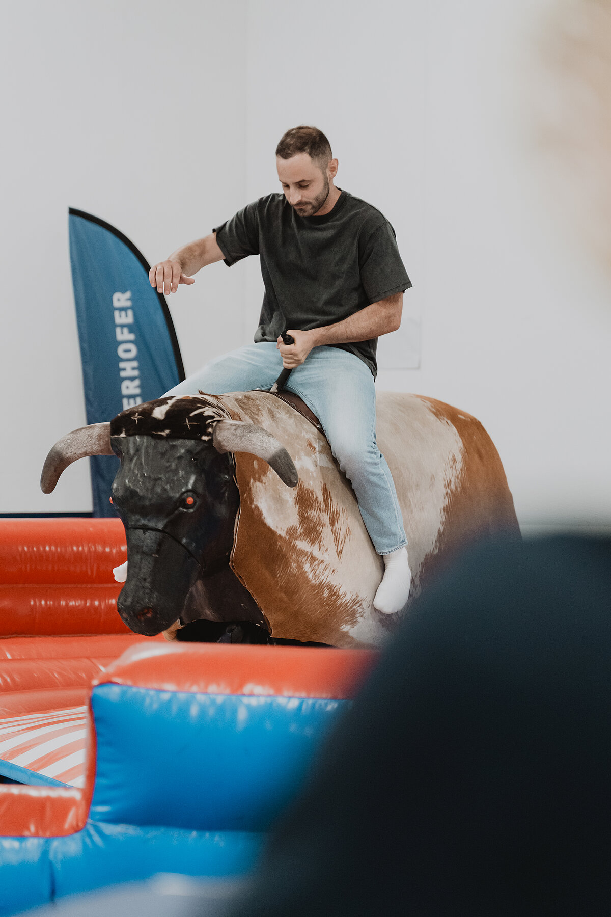 Ein Mann in Jeans und T-Shirt reitet auf einem mechanischen Bullen in einer luftgefüllten Arena. Der Bulle ist von rot-blauen Luftkissen umgeben. Im Hintergrund ist ein blauer Vertikalbanner zu sehen.