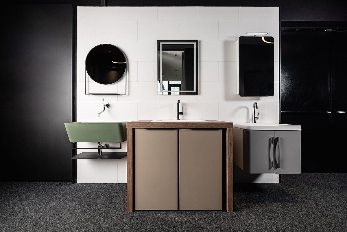 Minimalistisches Badezimmer-Design mit zwei modernen Waschbecken: ein grünes und ein weißes. Drei Spiegel an einer weißen Fliesenwand, darunter einer rund und zwei rechteckig. Ein Holzschrank steht zentral. Dezente und elegante Anordnung der Sanitäranlagen.