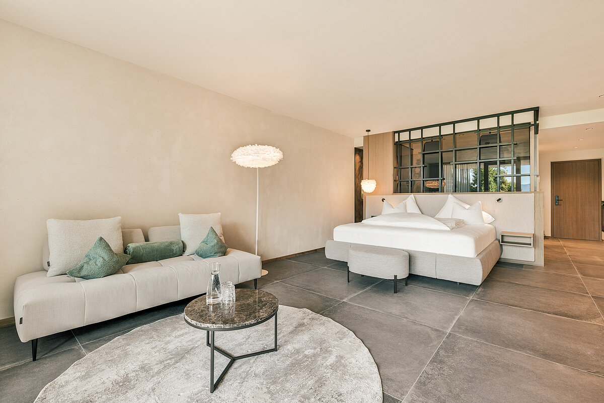 Modernes Hotelzimmer mit großem Doppelbett, grau gepolstertem Sofa, runden Beistelltisch und Stehlampe auf hellem Teppich. Eine Glaswand trennt den Schlafbereich. Dezente Farbtöne und minimalistisches Design prägen den Raum.