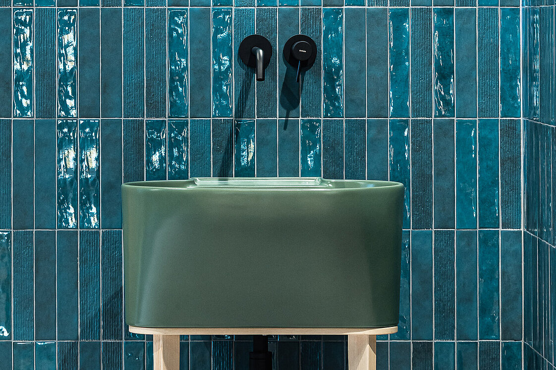 Ein Badezimmer mit auffälligen, vertikal verlegten blauen Fliesen. Ein rundes Spiegel hängt über einem modernen, grünen Waschbecken mit schwarzen Armaturen. Das Waschbecken steht auf einem hellen Holzgestell.