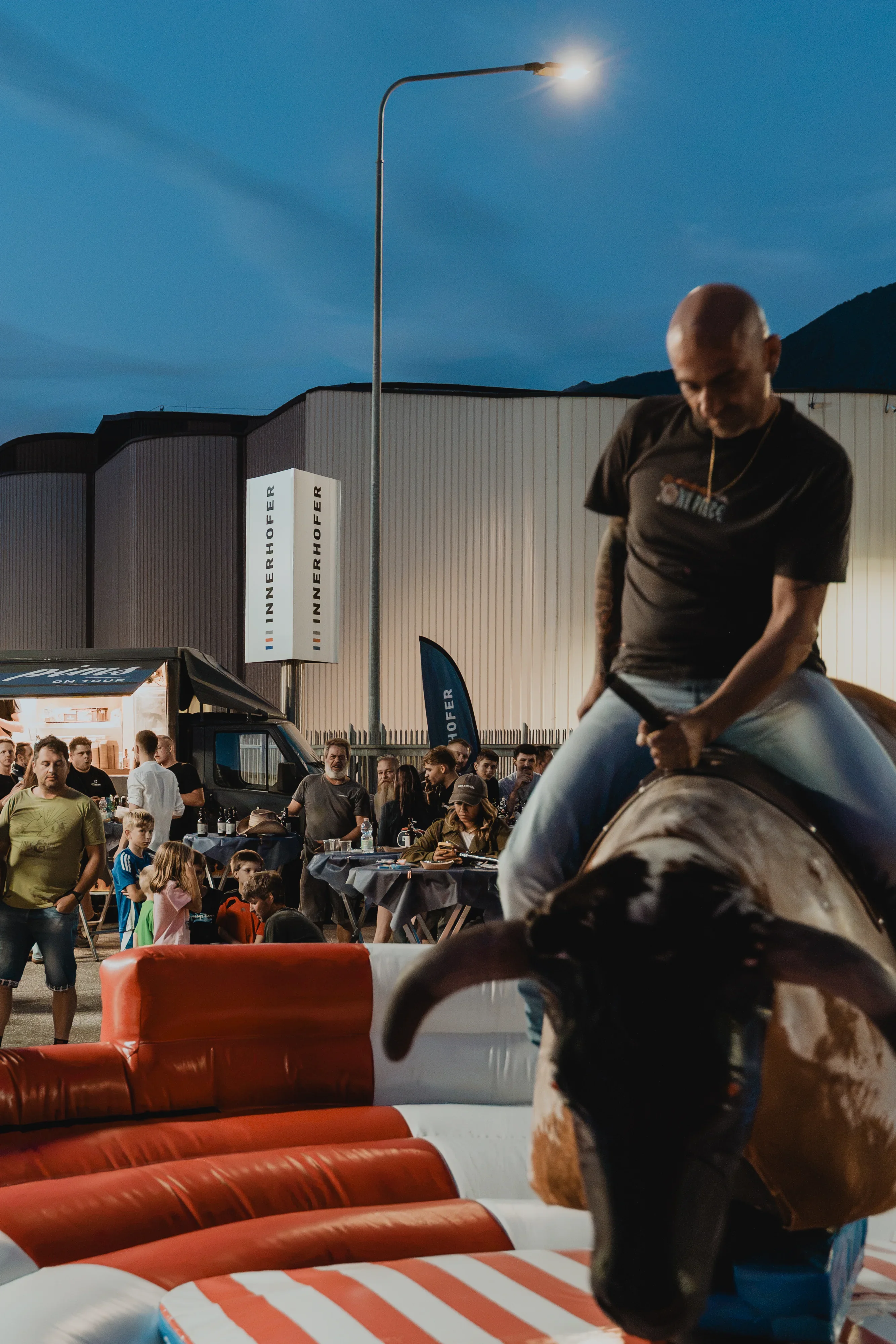 Ein Mann reitet auf einem mechanischen Bullen bei einer Veranstaltung im Freien. Das Publikum schaut zu, während im Hintergrund Abenddämmerung herrscht. Der Boden des Bullenarena ist in rot, weiß und blau gestaltet.