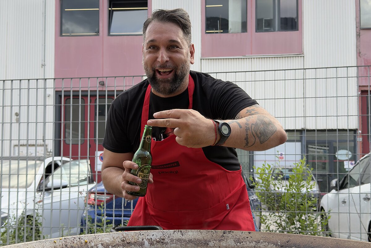 Ein Mann in roter Schürze steht lächelnd vor einem großen Paella-Pfanne im Freien. Er hält eine Flasche und scheint die Paella zuzubereiten. Im Hintergrund ist ein Gebäude mit rosa und weißen Fassaden zu sehen.