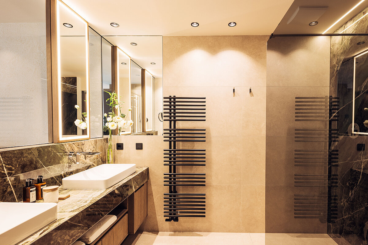 Modernes Badezimmer mit zwei Waschtischen und großen Spiegeln. Die Wände bestehen aus beigem Stein und dunklem Marmor. Ein schwarzer Handtuchheizkörper ist an der Wand montiert, und die Beleuchtung ist indirekt. Dekorative Pflanzen und Seifenspender sind auf der Theke platziert.