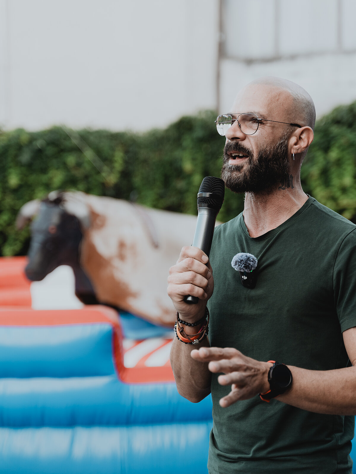 Ein Mann mit Brille und Bart hält ein Mikrofon und spricht. Er trägt ein dunkelgrünes T-Shirt. Im Hintergrund ist ein mechanischer Bulle auf einem bunten, gepolsterten Untergrund zu sehen. Die Szene findet draußen statt, mit einer Hecke im Hintergrund.