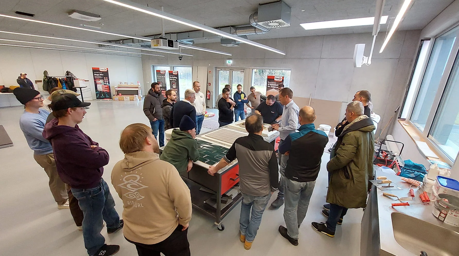 Eine Gruppe von Personen versammelt sich in einem Schulungsraum um einen Tisch mit Werkzeugen. Ein Dozent demonstriert eine Technik, während die Teilnehmer aufmerksam zusehen. Die Atmosphäre wirkt konzentriert und interessiert.