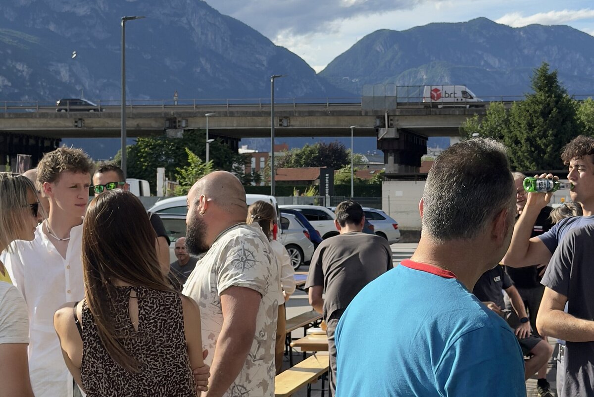 Eine Gruppe von Menschen steht im Freien und unterhält sich, mit einer Berglandschaft im Hintergrund. Der Himmel ist teilweise bewölkt, und es ist sonnig.