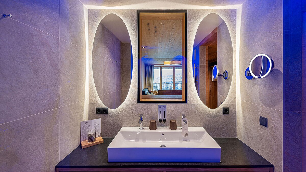 Elegantes Badezimmer mit zwei ovalen, beleuchteten Spiegeln über einem rechteckigen Waschbecken. Ein großer Wandspiegel reflektiert ein Fenster, das einen Blick ins Freie bietet. Die Beleuchtung erzeugt eine warme, moderne Atmosphäre.