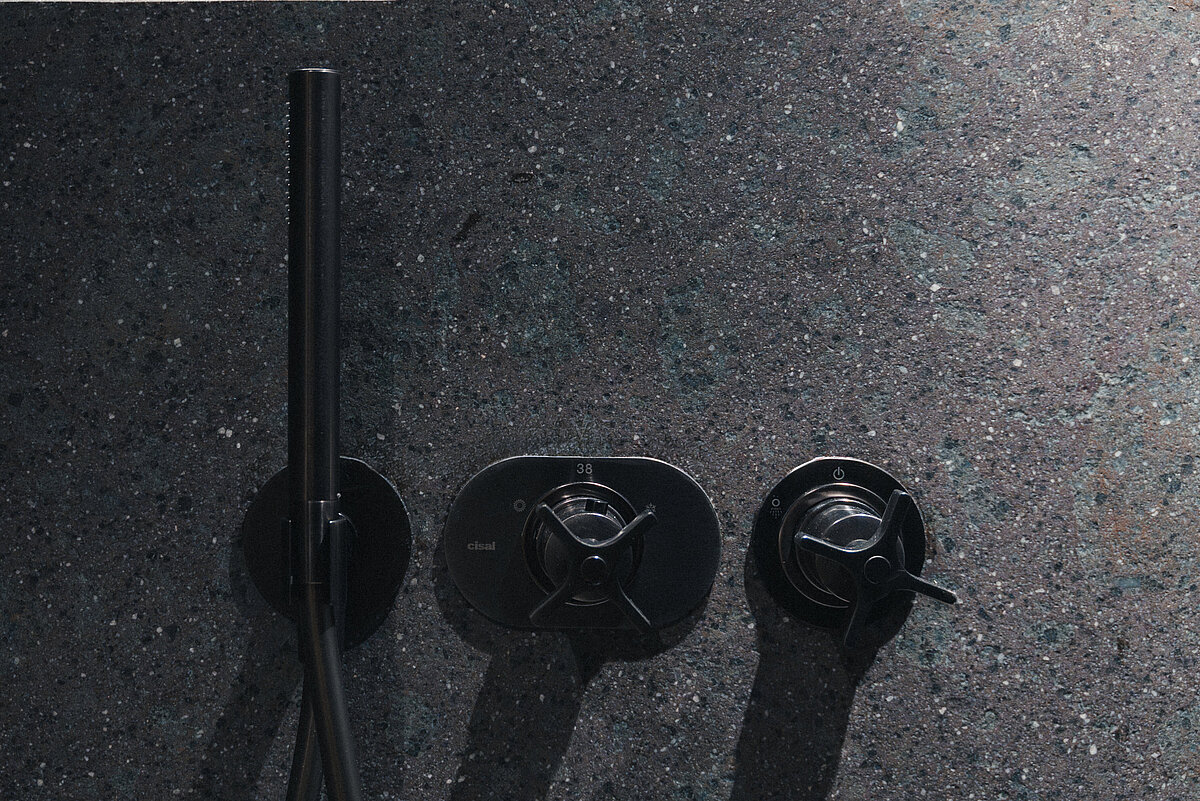 Schwarze Duscharmaturen und Schlauch an einer anthrazitfarbenen, strukturierten Wand mit großflächigen Fliesen.