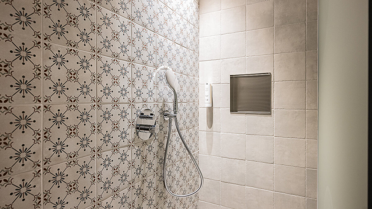 Moderne Dusche mit floral gemusterten Kacheln an der Wand. Eine Regendusche und eine Handbrause sind installiert. Ein kleines, eingebautes Regal in der Fliesenwand bietet Ablagefläche. Minimalistisches und gepflegtes Design.