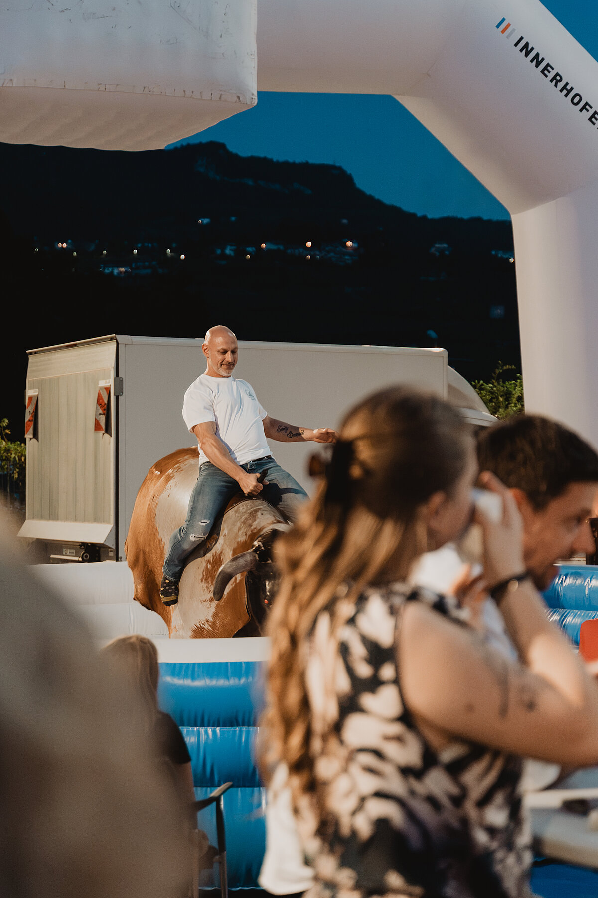 Ein Mann in einem weißen T-Shirt reitet auf einem mechanischen Bullen bei einem Outdoor-Event. Menschen im Vordergrund sehen ihm zu. Im Hintergrund sind Hügel und Gebäude im Abendlicht zu erkennen.
