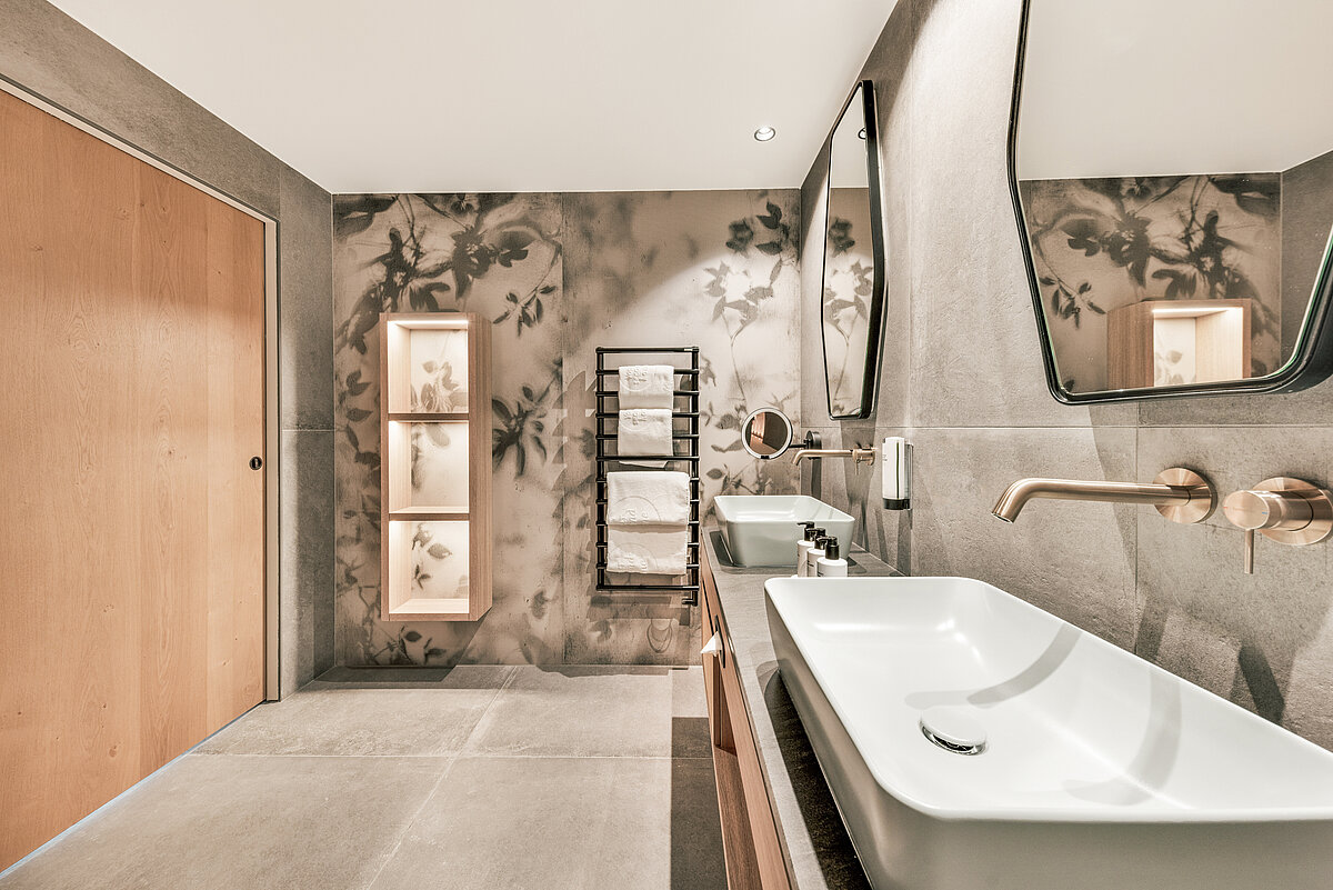 Modernes Badezimmer mit zwei rechteckigen Waschbecken, Wandhähnen, zwei großen Spiegeln und hölzernen Badschränken. Die Wände sind in einem grauen Design gehalten, mit einem floralen Muster an einer. Rechts ist ein Handtuchhalter, links eine Holztür.